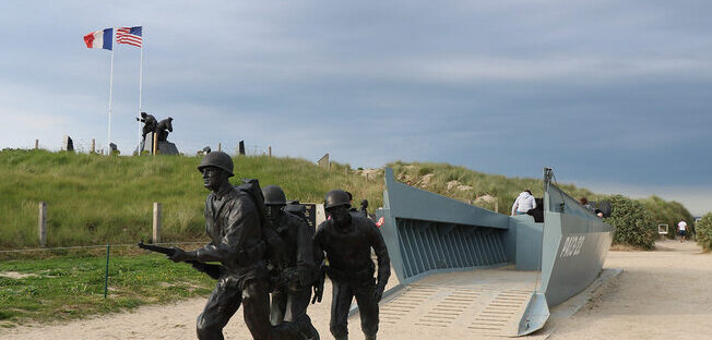 a picture of a statue and Land craft assault boat, several soldiers are marching out of the boat and libertaing france there are two flags in the background one french one american